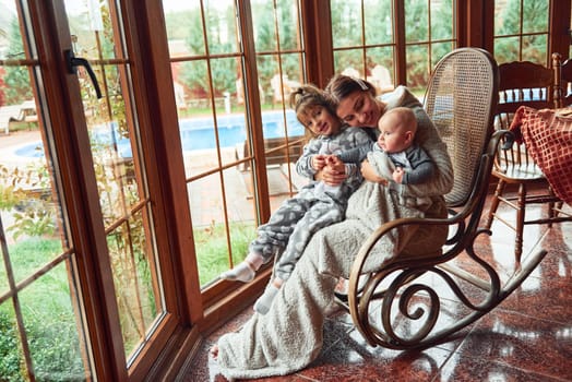 Happy mother sits with her little son and daughter on the chair near window.