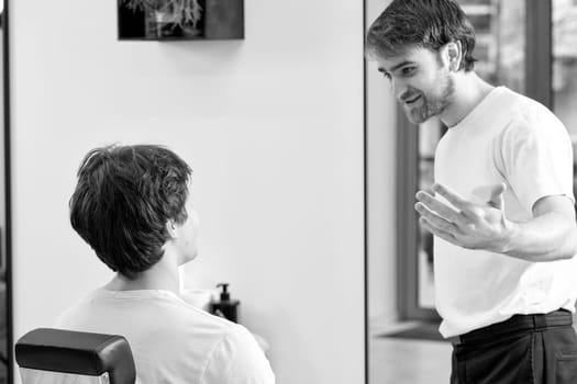 friendly barber talking to caucasian man while sitting in chair before haircut at barbershop