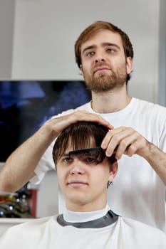 professional male barber combing caucasian man while sitting in chair before haircut at barbershop. client tells what haircut he wants