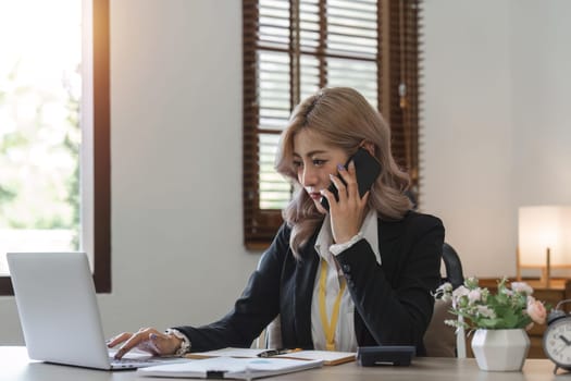 Happy young asian woman talking on the mobile phone and working smiling while sitting at her working place in office.