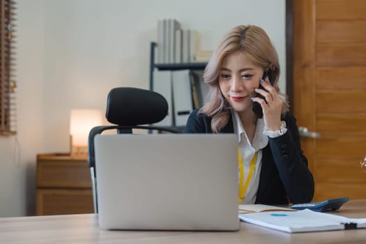 Happy young asian woman talking on the mobile phone and working smiling while sitting at her working place in office.