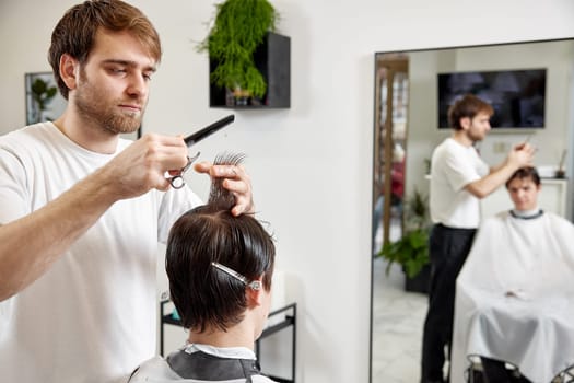 young caucasian man getting haircut by professional male hairstylist using comb and grooming scissors at modern white barber shop.