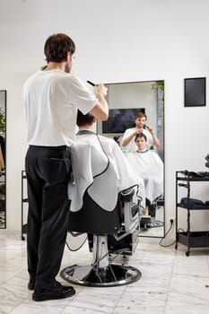 young caucasian man getting haircut by professional male hairstylist using comb and grooming scissors at modern white barber shop.