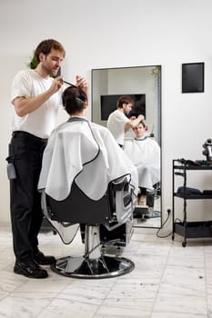 young caucasian man getting haircut by professional male hairstylist using comb and grooming scissors at modern white barber shop.