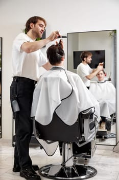 young caucasian man getting haircut by cheerful professional male hairstylist using comb and grooming scissors at modern white barber shop.