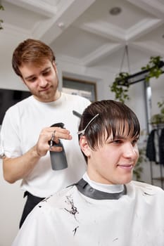 happy man getting haircut by professional male hairstylist using comb and sprayer at barber shop.