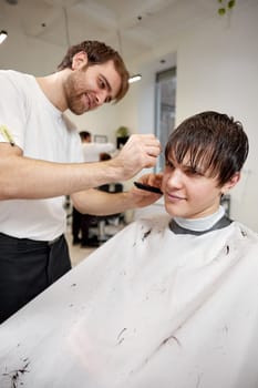 young caucasian man getting haircut by professional male hairstylist using comb and grooming scissors at barber shop.