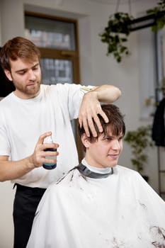 young caucasian man getting haircut by professional male hairstylist using sprayer at barber shop.