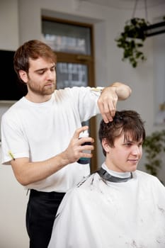 young caucasian man getting haircut by professional male hairstylist using sprayer at barber shop.