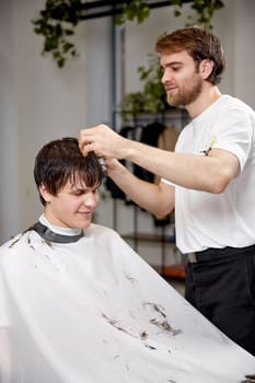 young caucasian man getting haircut by professional male hairstylist using sprayer at barber shop.