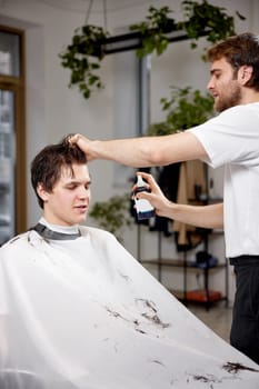 young caucasian man getting haircut by professional male hairstylist using sprayer at barber shop.