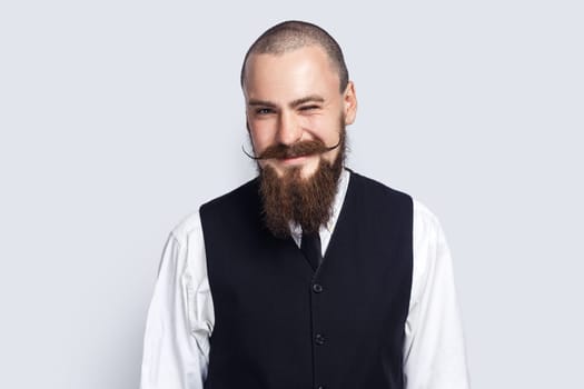 Portrait of positive happy handsome man with beard and mustache standing and winking to camera, flirting with somebody, smiling happily. Indoor studio shot isolated on gray background.