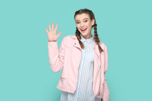 Portrait of friendly lovely teenager girl with braids wearing pink jacket waving hand, saying hello or bye, looking at camera with happiness. Indoor studio shot isolated on green background.