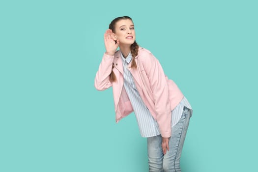 Portrait of curious young teenager girl with braids wearing pink jacket standing with palm near ear, trying to listen secret. Indoor studio shot isolated on green background.