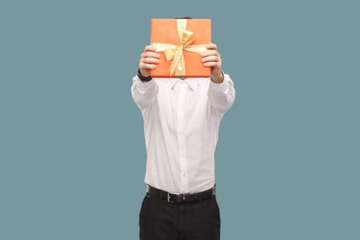 Portrait of unknown anonymous man holding out and covering his face with red gift box, giving present for anniversary, wearing Indoor studio shot isolated on light blue background.