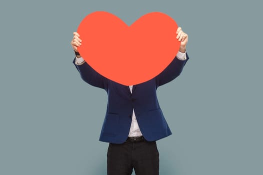 Unknown man standing hiding his face behind big red heart, being shy to say about his feelings, wearing official style suit. Indoor studio shot isolated on light blue background.