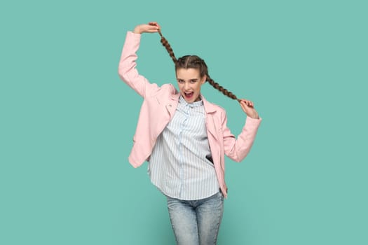 Portrait of excited amazed teenager girl with braids wearing pink jacket having fun, pulling her pigtails, looking at camera with smile. Indoor studio shot isolated on green background.