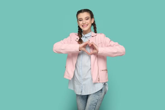 Portrait of smiling positive happy teenager girl with braids wearing pink jacket standing showing heart shape with hands, looking at camera. Indoor studio shot isolated on green background.