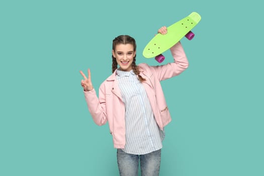 Portrait of pretty teenager girl with braids wearing pink jacket holding penny skateboard, spending free time in active way, showing v sign. Indoor studio shot isolated on green background.