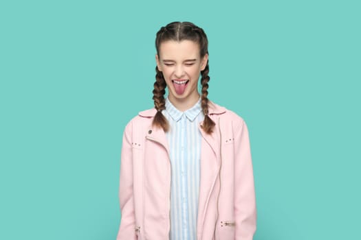 Portrait of funny childish teenager girl with braids wearing pink jacket standing with closed eyes, sticking her tongue out, having fun. Indoor studio shot isolated on green background.