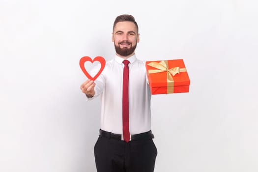 Portrait of joyful young adult handsome man in white shirt standing holding present box and red heart, gift for Valentines Day. Indoor studio shot isolated on gray background.