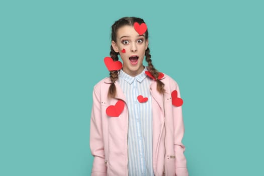 Portrait of amazed surprised teenager girl with braids wearing pink jacket standing covered with little red hears, looking at camera with open mouth. Indoor studio shot isolated on green background.