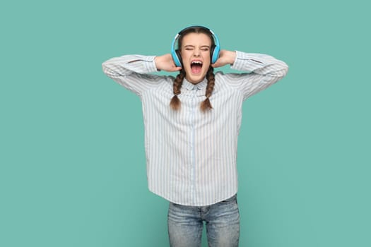 Excited crazy teenager girl with braids wearing striped shirt standing listening favorite music with headphones, singing with widely ope mouth. Indoor studio shot isolated on green background.