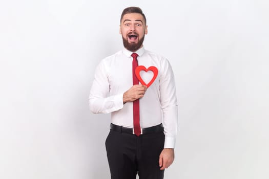 Portrait of extremely happy excited handsome man in white shirt holding red heart, looking at camera and screaming, falling in love. Indoor studio shot isolated on gray background.