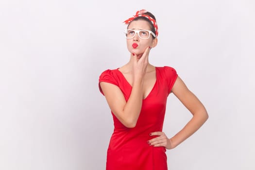 Portrait of thoughtful beautiful woman in red dress and head band standing holding her chin, looking away, thinking, remembering important information. Indoor studio shot isolated on gray background.