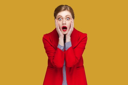 Portrait of amazed surprised woman with red lips with hands on cheeks, looking at camera with big eyes, sees something scary, wearing red jacket. Indoor studio shot isolated on yellow background.