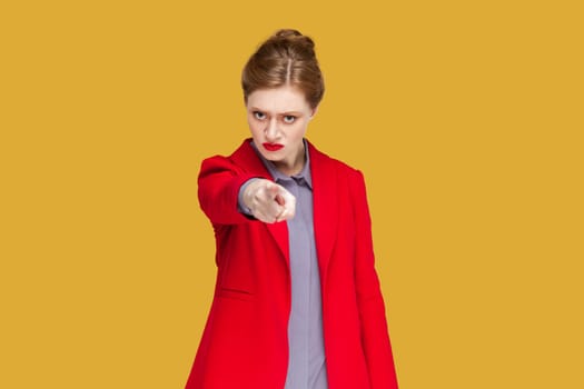 Portrait of strict serious woman with red lips standing indicating index finger to you, looking at camera with bossy expression, wearing red jacket. Indoor studio shot isolated on yellow background.