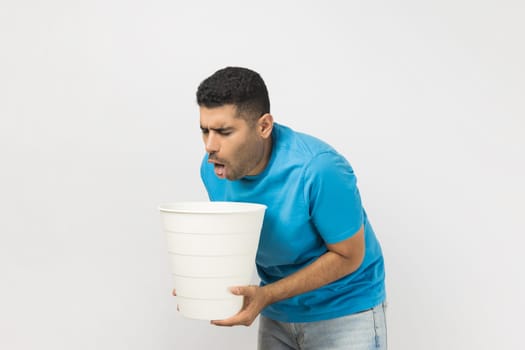 Portrait of sick unhealthy ill unshaven man wearing blue T- shirt standing suffering stomachache, feels nausea and vomits, holding bin in hands. Indoor studio shot isolated on gray background.