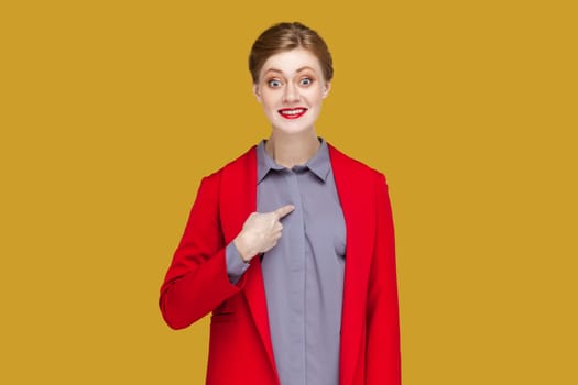 Portrait of smiling proud woman with red lips standing pointing finger at herself with happy facial expression, being in good mood, wearing red jacket. Indoor studio shot isolated on yellow background