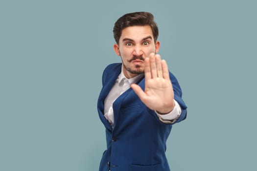 Portrait of strict bossy man with mustache standing showing palm, stop gesture, expressing negative emotions, wearing white shirt and jacket. Indoor studio shot isolated on light blue background.