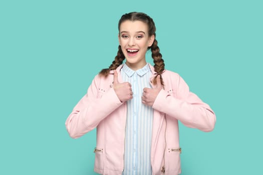 Portrait of excited cheerful smiling teenager girl with braids wearing pink jacket standing showing thumbs up, saying she likes party. Indoor studio shot isolated on green background.