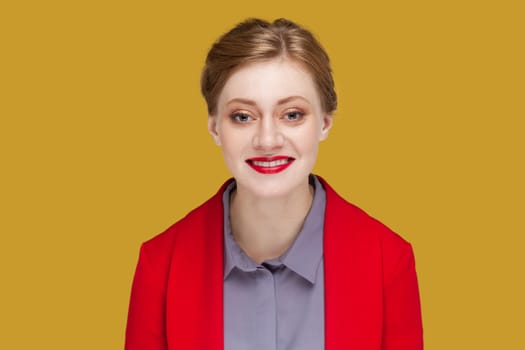 Portrait of happy optimistic woman with red lips standing looking at camera with toothy smile, expressing happiness, wearing red jacket. Indoor studio shot isolated on yellow background.