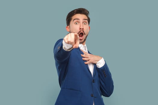 Astonished shocked man with mustache standing pointing finger to camera, keeps mouth widely open, looks scared, wearing white shirt and jacket. Indoor studio shot isolated on light blue background.