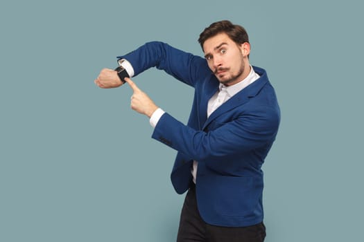 Portrait of serious assertive man with mustache standing pointing at his wristwatch, saying you have no time, wearing white shirt and jacket. Indoor studio shot isolated on light blue background.