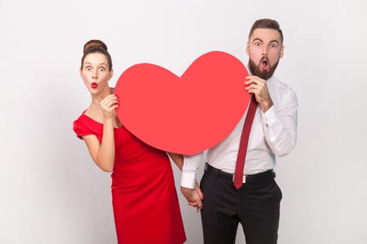 Portrait of amazed shocked man in white shirt and woman in red dress standing together, peeping out from big heart, expressing love. Indoor studio shot isolated on gray background.