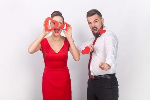 Portrait of crazy man in white shirt and amazed woman in red dress standing together, holding heart figures, being emotive. Indoor studio shot isolated on gray background.