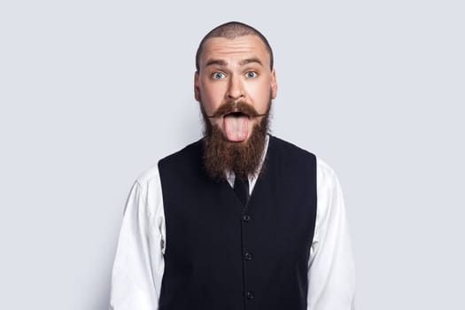 Portrait of childish impolite man with beard and mustache showing tongue out, demonstrating bad manners, looking at camera with sticking tongue. Indoor studio shot isolated on gray background.