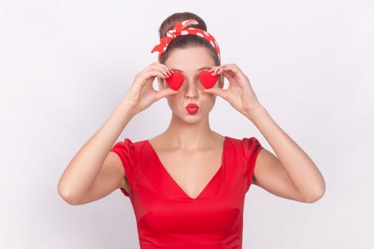 Portrait of romantic woman in red dress and head band covering her eyes with little red hearts, symbol of love, sending air kissing, keeps lips pout. Indoor studio shot isolated on gray background.