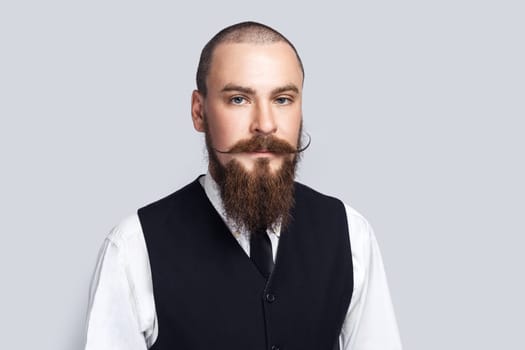 Portrait of serious attentive man with beard and mustache standing looking at camera with strict facial expression, being in bad mood. Indoor studio shot isolated on gray background.