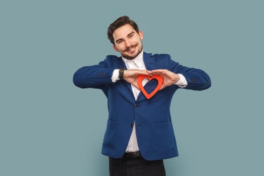 Romantic handsome man with mustache standing and showing red heart shaped figure, looking at camera with smile, wearing official style suit. Indoor studio shot isolated on light blue background.