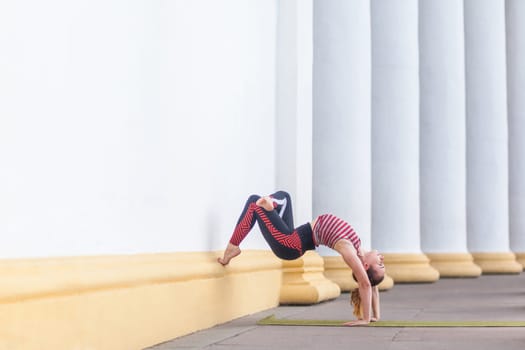 Full length side view of athletic beautiful young adult woman wearing sportswear doing wall-assisted handstand pose, practicing yoga, doing pilates or fitness sport exercise outdoor.