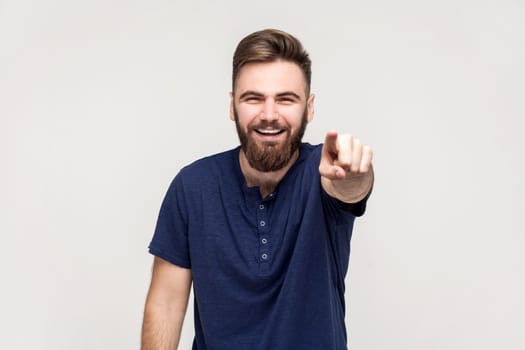 Portrait of amused handsome man with beard wearing dark blue T-shirt laughing out loud, pointing finger to camera, indicating ridiculous idiot. Indoor shot isolated on gray background.