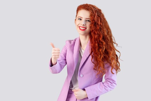 Portrait of satisfied delighted ginger businesswoman showing like gesture, recommend service, posing with thumb up, wearing lilac jacket. Indoor studio shot isolated on gray background.