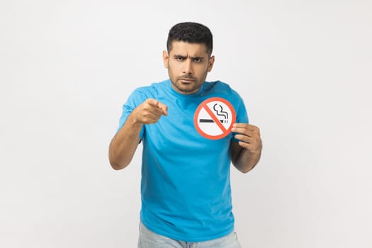 Portrait of serious unshaven man wearing blue T- shirt standing showing no smoking sign, pointing at camera, warning you about danger for your health. Indoor studio shot isolated on gray background.