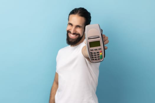 Portrait of cheerful man with beard wearing white T-shirt holding out showing pos terminal, suggesting you to use contactless payments. Indoor studio shot isolated on blue background.