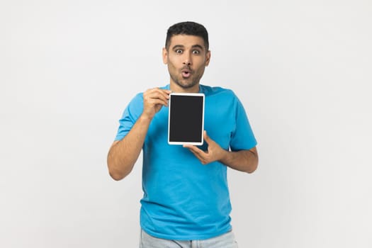 Portrait of astonished shocked unshaven man wearing blue T- shirt standing showing table t with black empty display, copy space for advertisement. Indoor studio shot isolated on gray background.